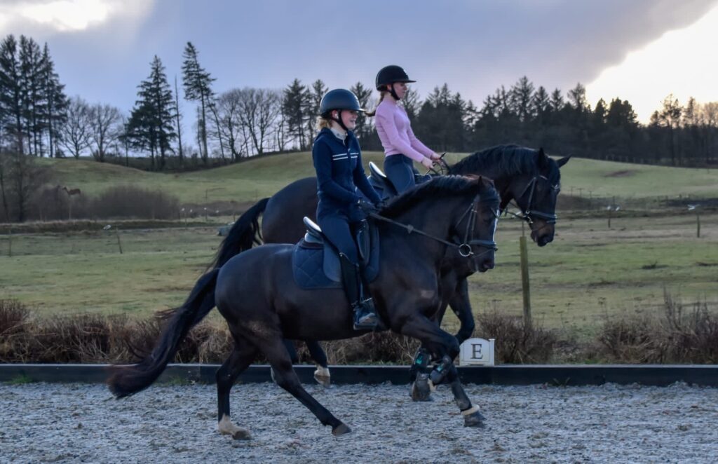 2 unge piger der træner dressur på den udendørs ridebane med foldene i baggrunden